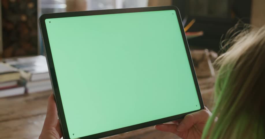 A person in a room looking at a tablet with a table and books in the background