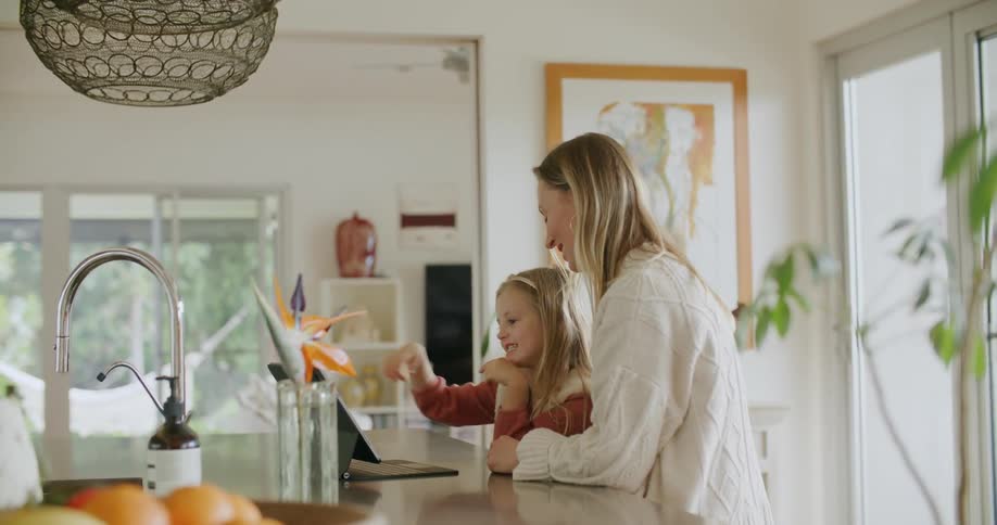 A woman and a girl in the kitchen looking at a laptop with a wall and doorway in the background