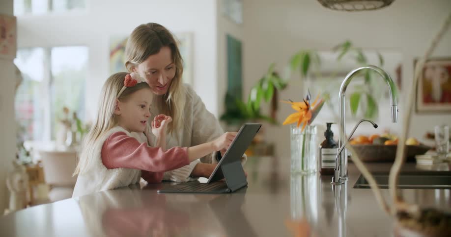 Mother smiles and talks while watching daughter work on tablet as they sit at kitchen island