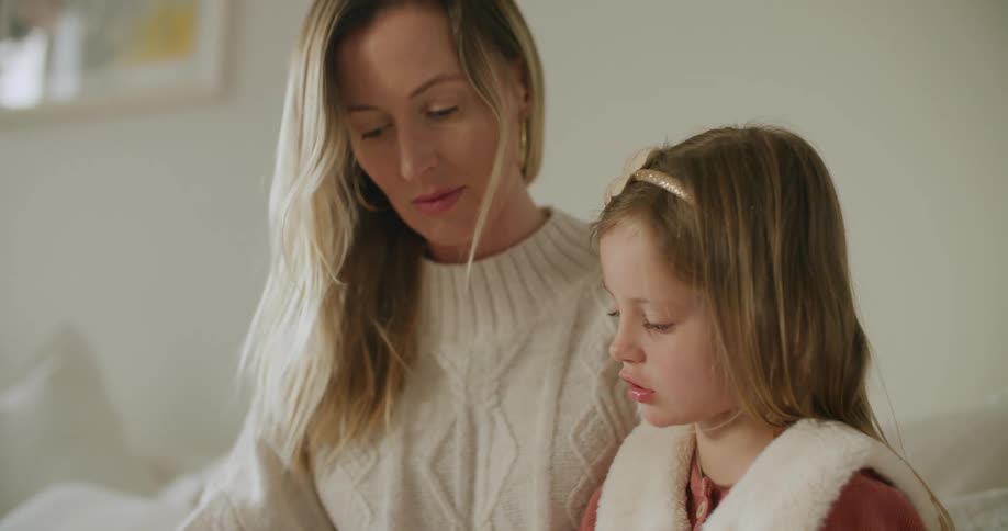 Blonde mother helps daughter use tablet sitting on couch at home