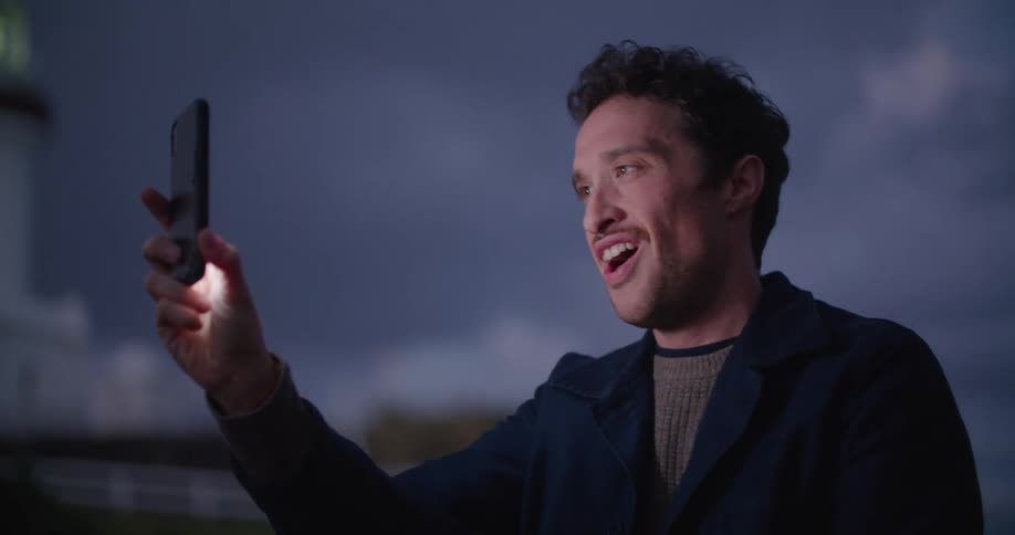 Man stands outside at night near lighthouse talking and gesturing while video calling on mobile phone