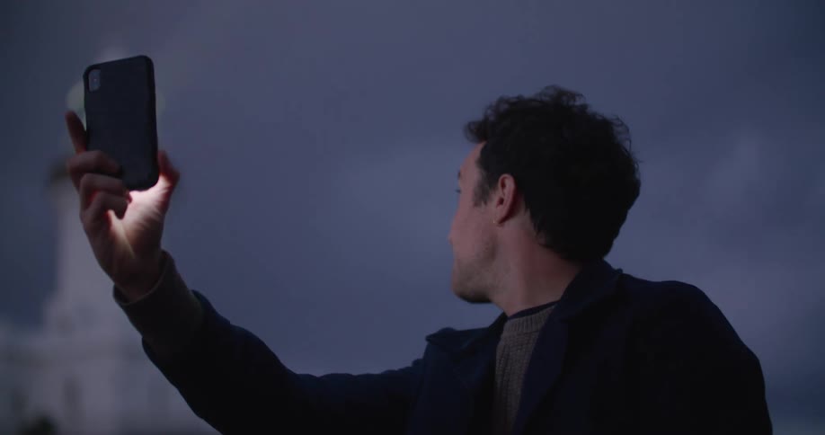 Man stands outside talking via video call on mobile phone moving phone to show nearby scenic lighthouse