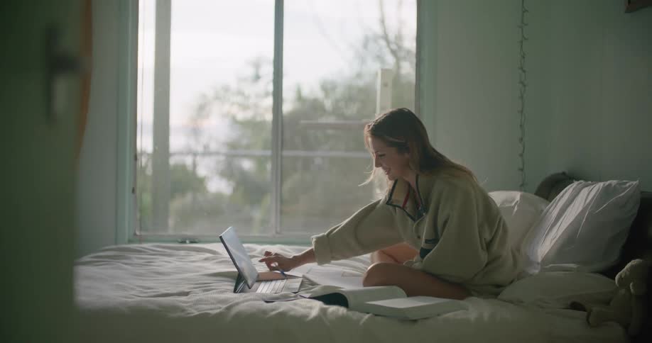 A woman sits on a bed having a video call on a laptop in front of her