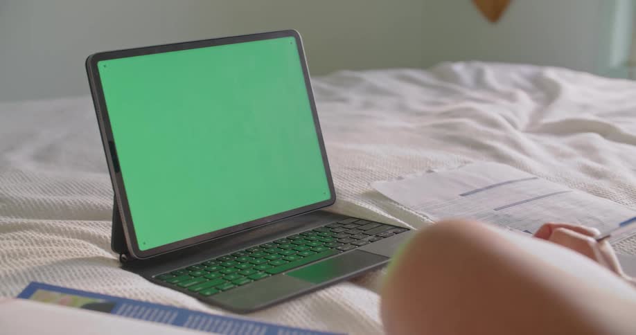 Bare legged person sits on bed near tablet writing and flipping through textbook