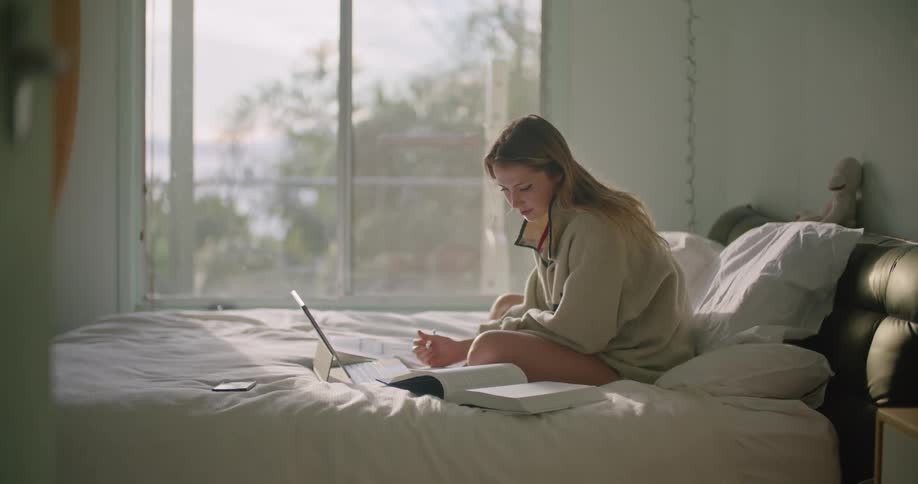 A woman in a room sitting on a bed with a laptop writing in a book with a pen