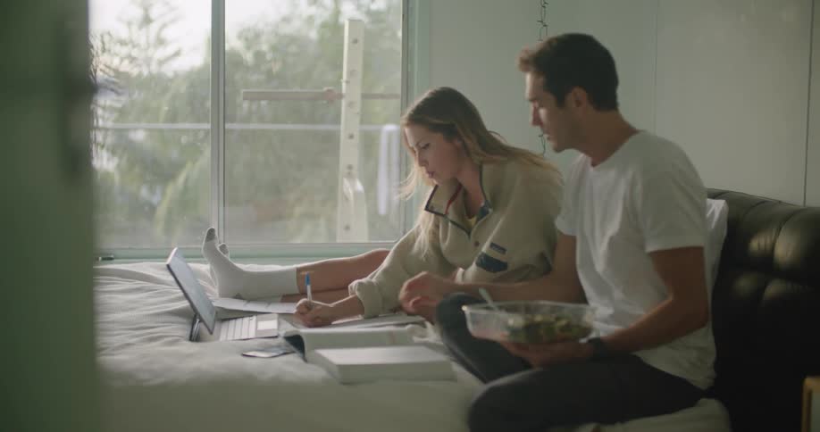 A man eats a salad while sitting in bed and helping his girlfriend study on a laptop