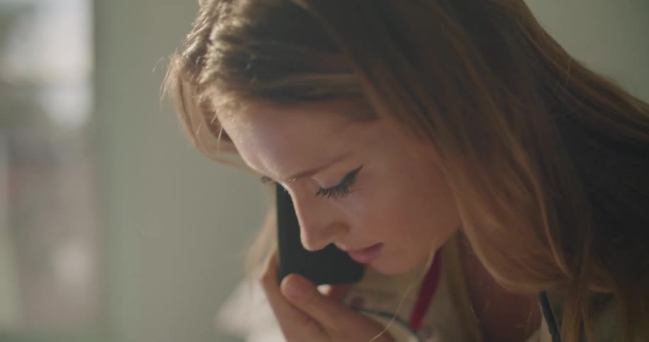Young blonde woman sitting in bed at home talking on the phone while taking notes with pen and paper