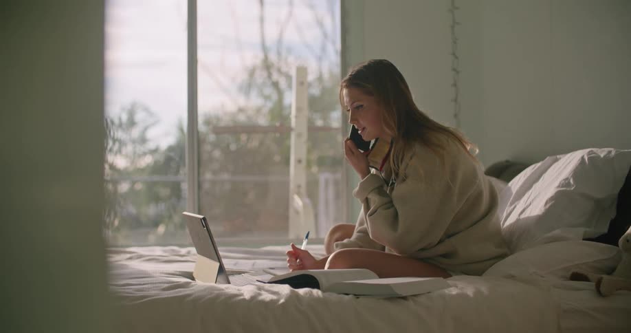 Blonde woman with oversized sweatshirt sits on bed an uses tablet while talking on smartphone
