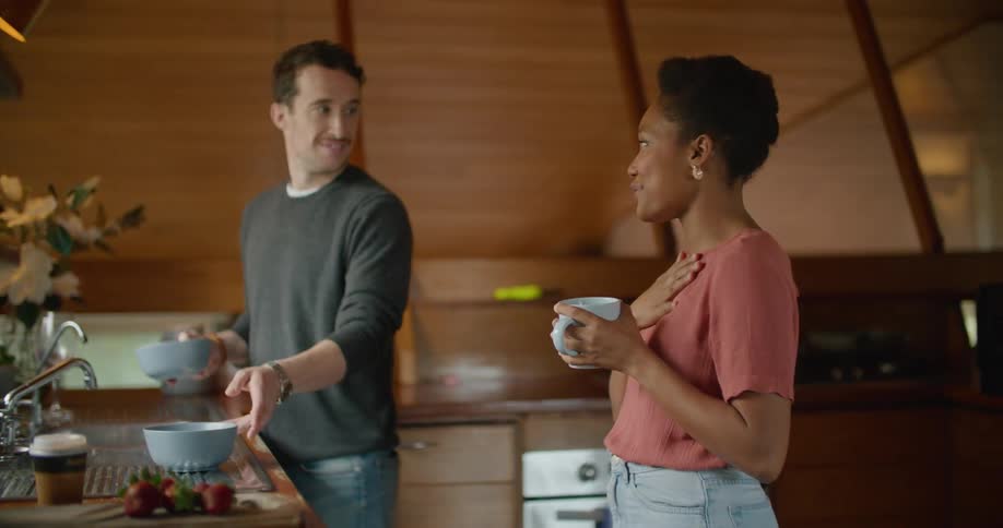 A man picks up two bowls of food while chatting with his girlfriend in their kitchen