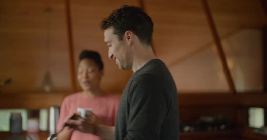 A man and woman stand in the kitchen drinking from coffee mugs and laughing
