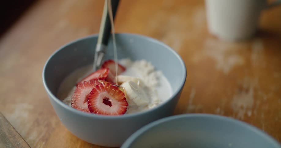 A spoon of honey is being added onto sliced strawberries and bananas in bowl