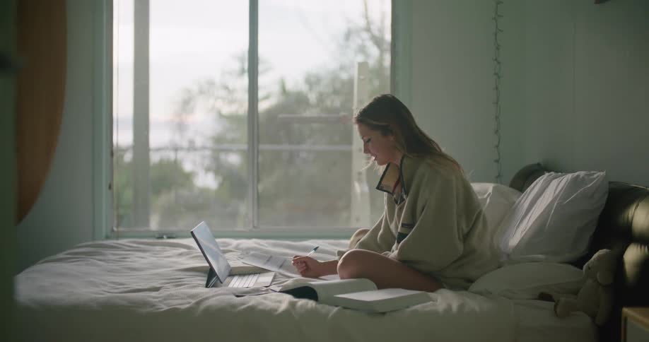 A teenage girl sits on her bed and does homework while video chatting on her laptop