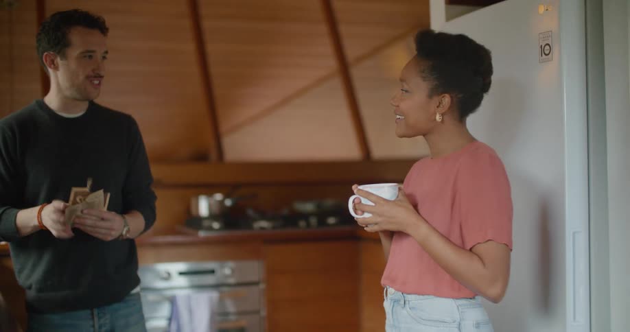 A woman holding a cup of coffee in a kitchen talks to a man as he makes oatmeal
