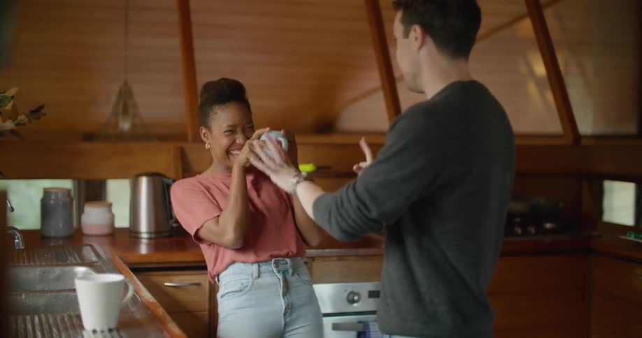 A man and woman stand in a kitchen talking and laughing while the woman holds a coffee mug
