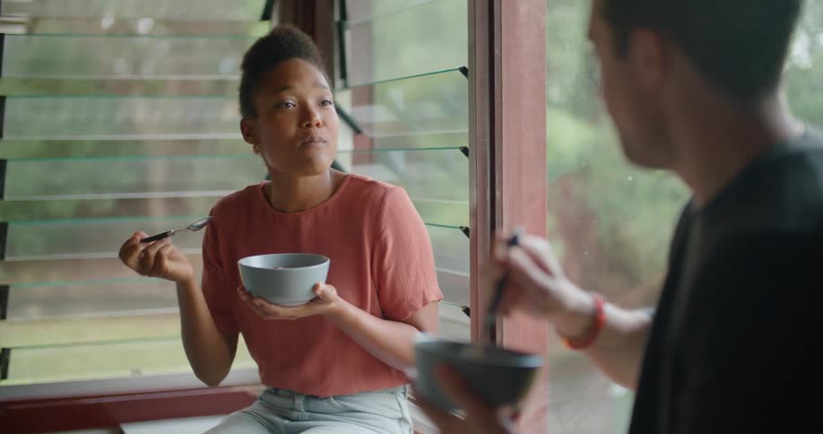 Man and woman sit near windows holding bowls and talking while they eat