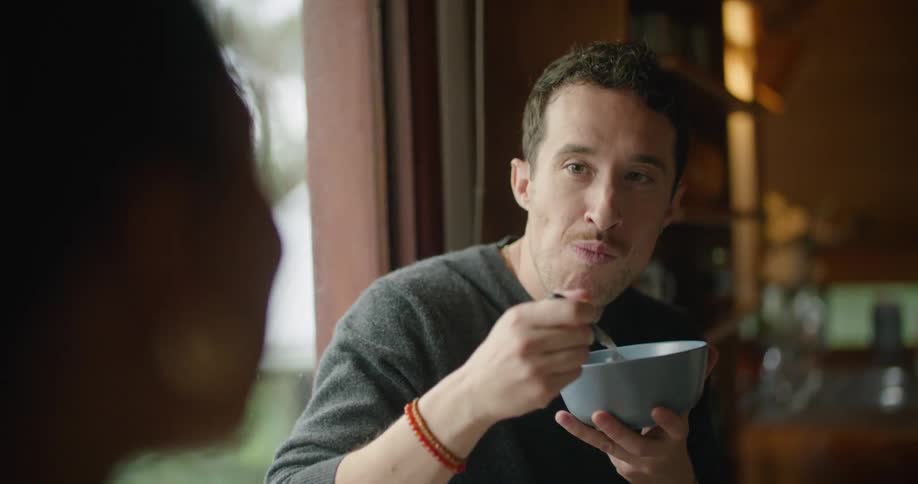 A man sits in a house and eats as he talks with a woman