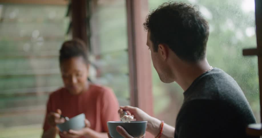 A man and woman sit on a window ledge eating from bowls