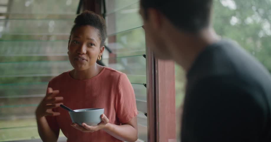 A man and woman sit by a window in a house and eat and talk