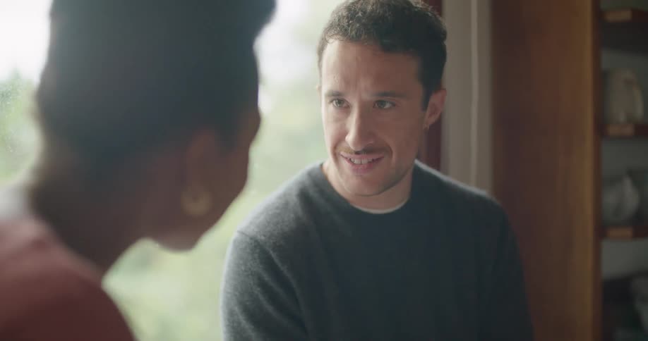 Man wearing grey sweatshirt and jeans sits on window pane and uses tablet with woman with short hair and red shirt