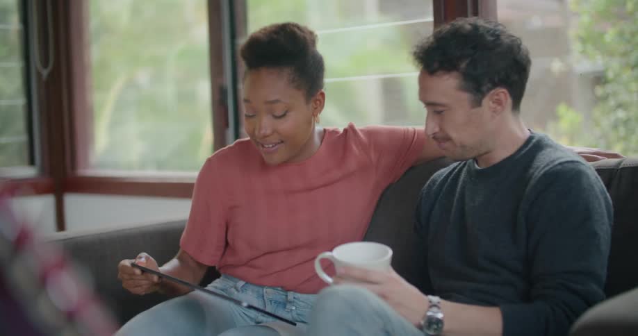 A woman talks to a man and shows him a tablet as she moves next to him on a sofa in a house during the day