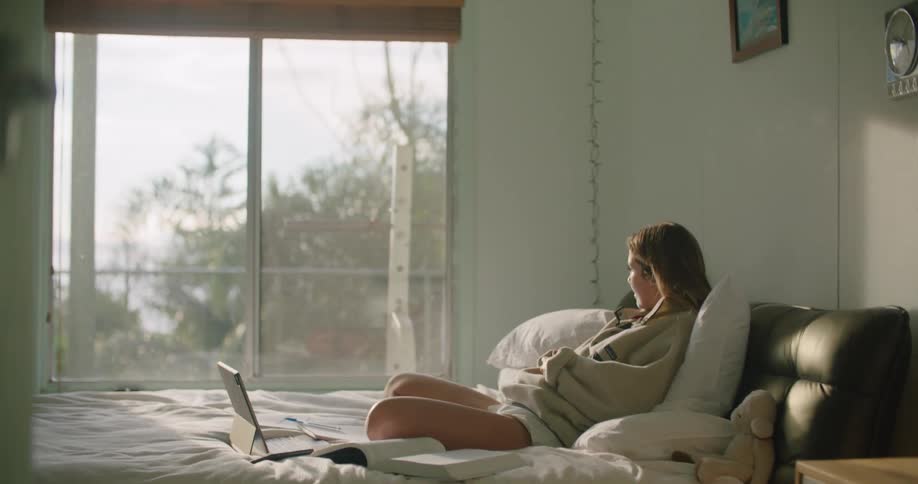 A woman sits on a bed with a laptop writing in a book then leans back on the pillows behind her