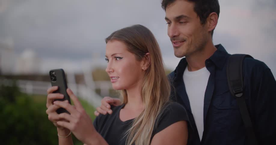 A man and a woman looking at a smart phone with a fence the sky and clouds in the background