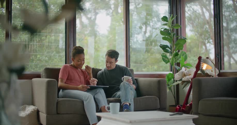 A man and woman sit side by side on a love seat smiling as they scroll on a tablet