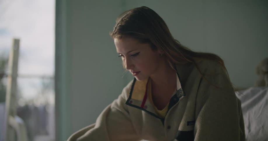 Woman sits on bed near open textbook using tablet and taking notes