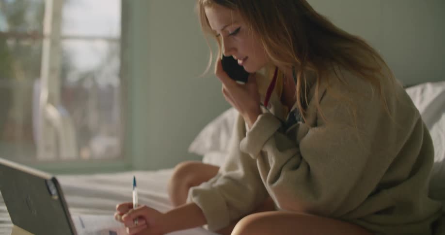 A woman gets help with homework on the phone while sitting in bed with her laptop and books