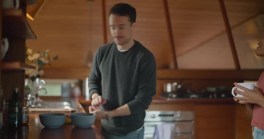 A woman holding a coffee mug talks to a man as he cleans a kitchen counter