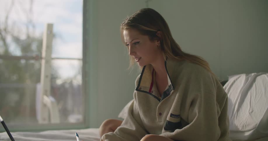 Blonde woman with oversize sweatshirt sitting on bed writes in notebook while using tablet