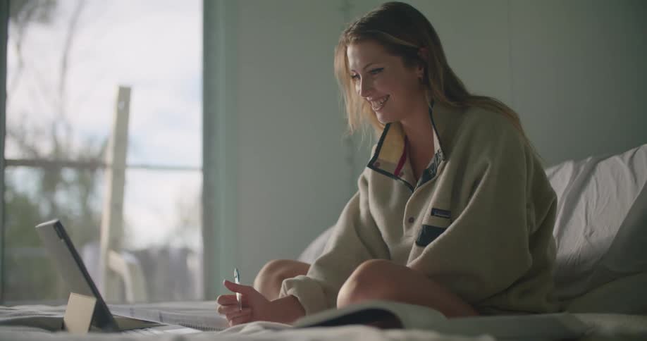 A woman in a bedroom sitting in a bed looking at a laptop while holding a pen