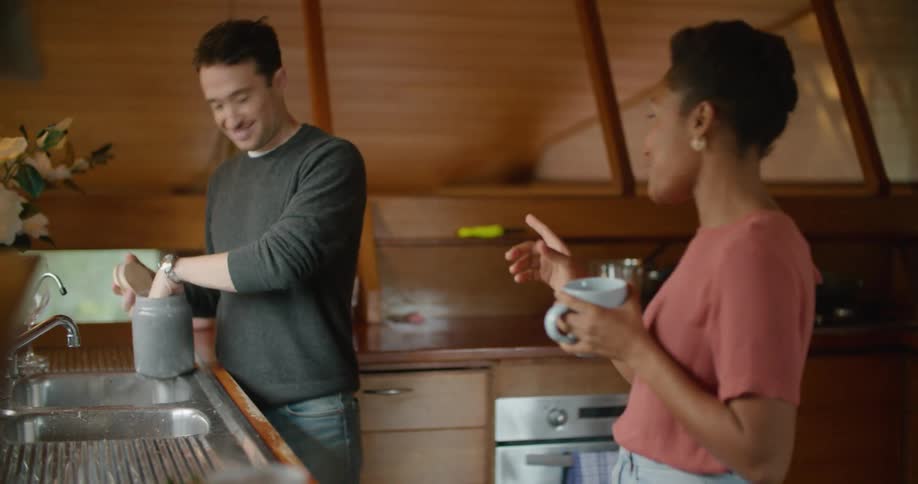 A woman holds a cup of coffee and talks and laughs with a man in a house