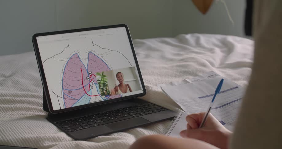 A person in a bedroom sitting in a bed looking at a laptop while writing on a notebook with a pen