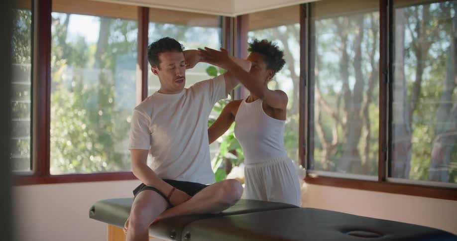 A man and a woman in a room with the man sitting on a massage table when a plant and windows in the background