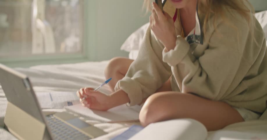 A woman sits on a bed talking on the phone with a laptop in front of her and writing in a notebook