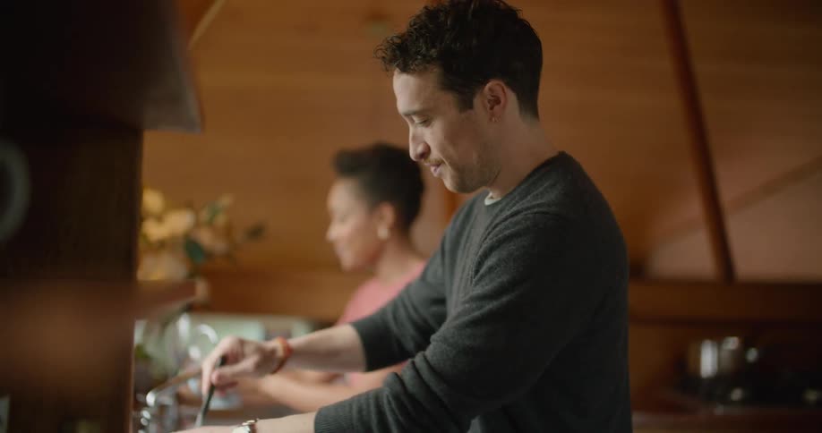 A man uses a spoon to stir two bowls next to strawberries on a cutting board while a woman drinks water in a mug