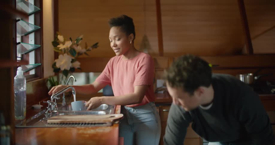 Couple wearing sweaters work together to wash dishes in wood paneled kitchen
