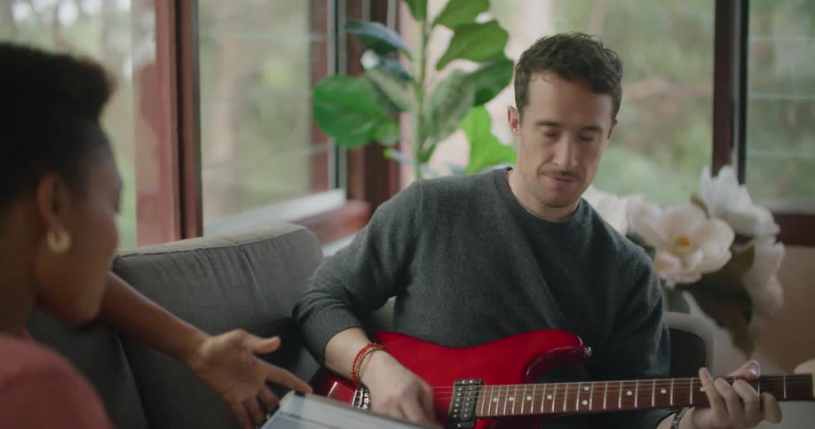 A man plays a guitar while speaking to a woman using a tablet next to him on a sofa in a house during the day