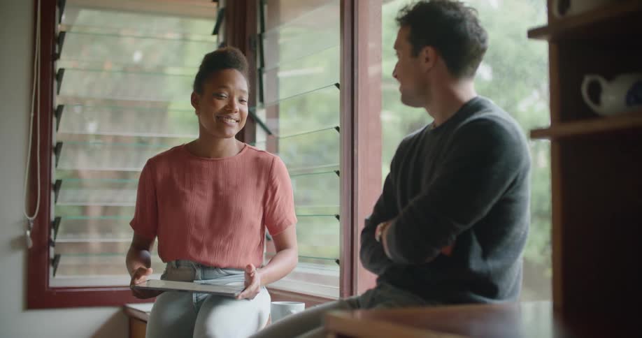 A couple sit and lean on their kitchen counter chatting while the woman uses a tablet