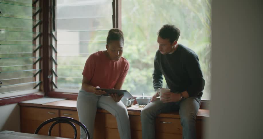 Man and woman sit near window sharing tablet with food on furniture between them