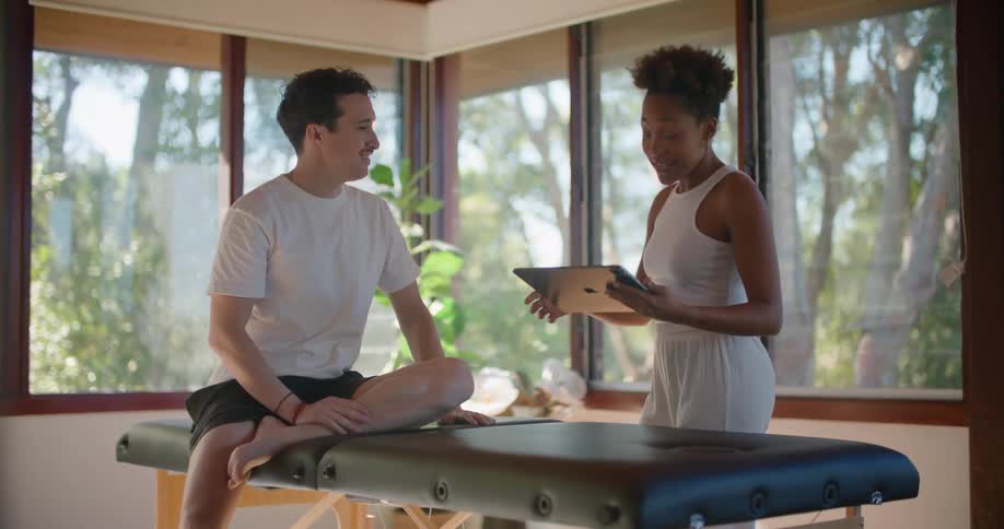 A man sits on a massage table while a woman holding a tablet talks to him