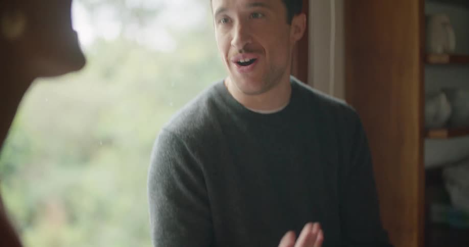 Man wearing sweater sits near window discussing image on tablet with woman