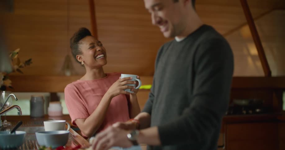 A man cuts a banana on a cutting board while talking to a woman as they stand together in a home kitchen during the day