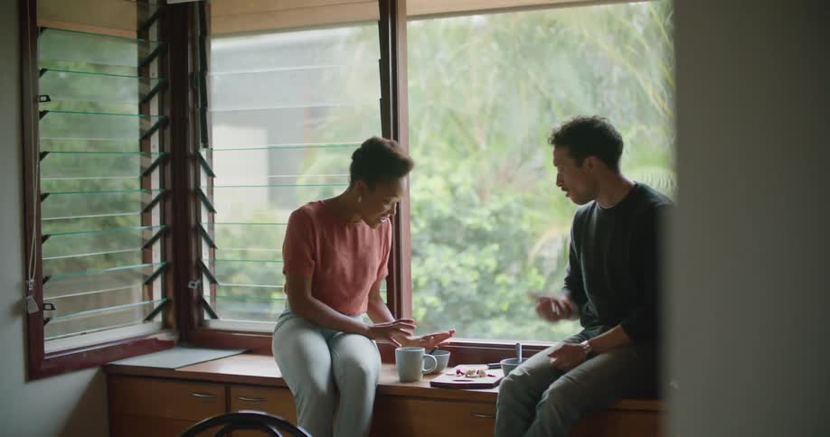 A man and woman sit by a window in a home and talk and eat