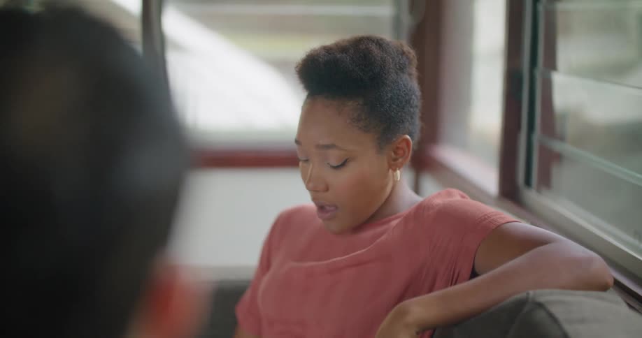Woman with short hair red t shirt and jeans sits on couch and uses tablet talking to man
