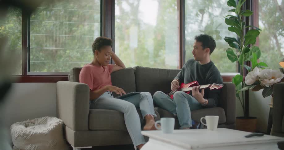 A man and a woman in a room sitting on a couch with a man holding electric guitar and the woman looking at a tablet