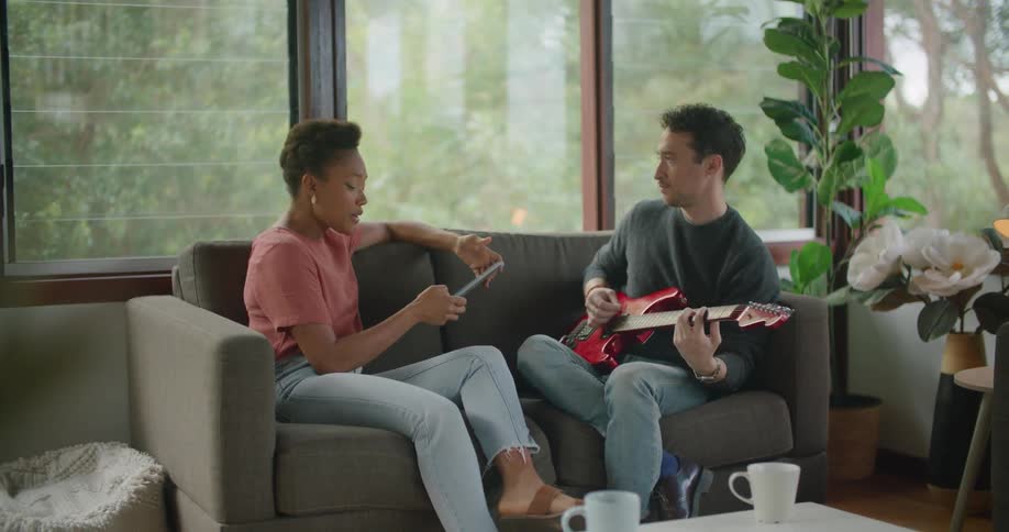 Man with grey sweater and jeans plays red electric guitar while woman with short hair red t shirt and jeans using tablet sitting on couch