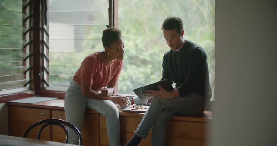 A man holds a tablet showing it to a woman sitting beside him on the top of a cabinet