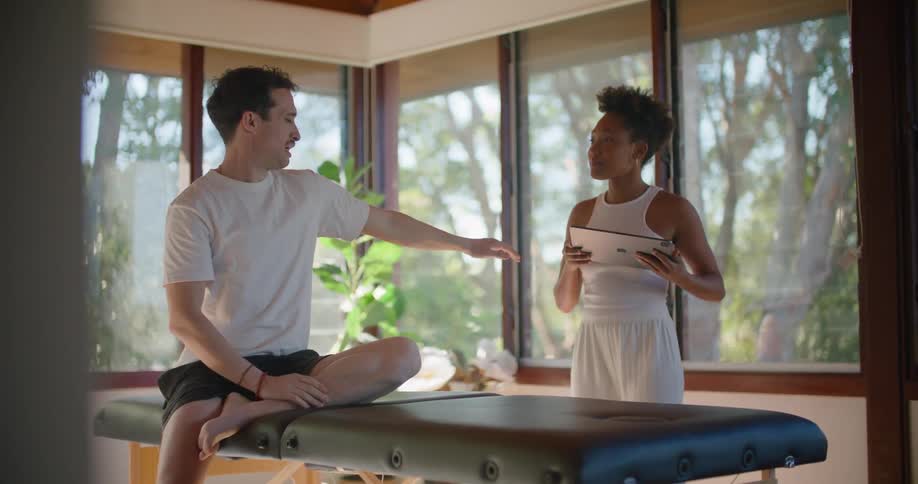 A man sits in a clinic and explains his pains to a physical therapist who then shows him information on a tablet